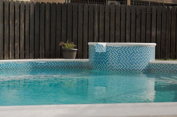 A swimming pool with a mosaic tile edge, a wooden fence in the background, and a potted plant beside the pool.