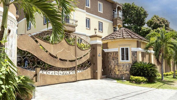 A two-story building with a gated entrance, a small attached structure with a tiled roof, and a well-maintained garden with palm trees. - Barri Vacation Home (Kingston)