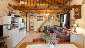 A cozy living room with a wooden ceiling, a sofa with plaid cushions, a dining table with a vase of dried flowers, and a kitchen area with a washing machine. - Holiday house Jano in Makarska, jacuzzi (Makarska)