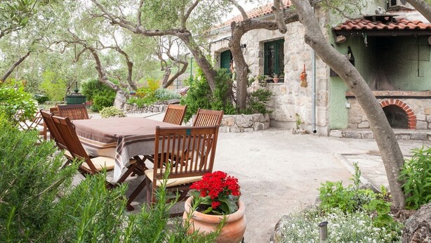 An outdoor dining area with a table and chairs, surrounded by greenery and a stone building with a tiled roof. - Holiday house Jano in Makarska, jacuzzi (Makarska)