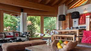 A cozy living room with a wooden ceiling, large windows, a dining area with a wooden table, and a fruit bowl on the table.