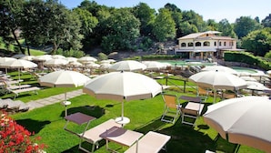 Un espace extérieur aménagé avec goût, doté de nombreuses parasols blancs et de chaises longues, entouré d’une végétation luxuriante et avec un grand bâtiment en arrière-plan.