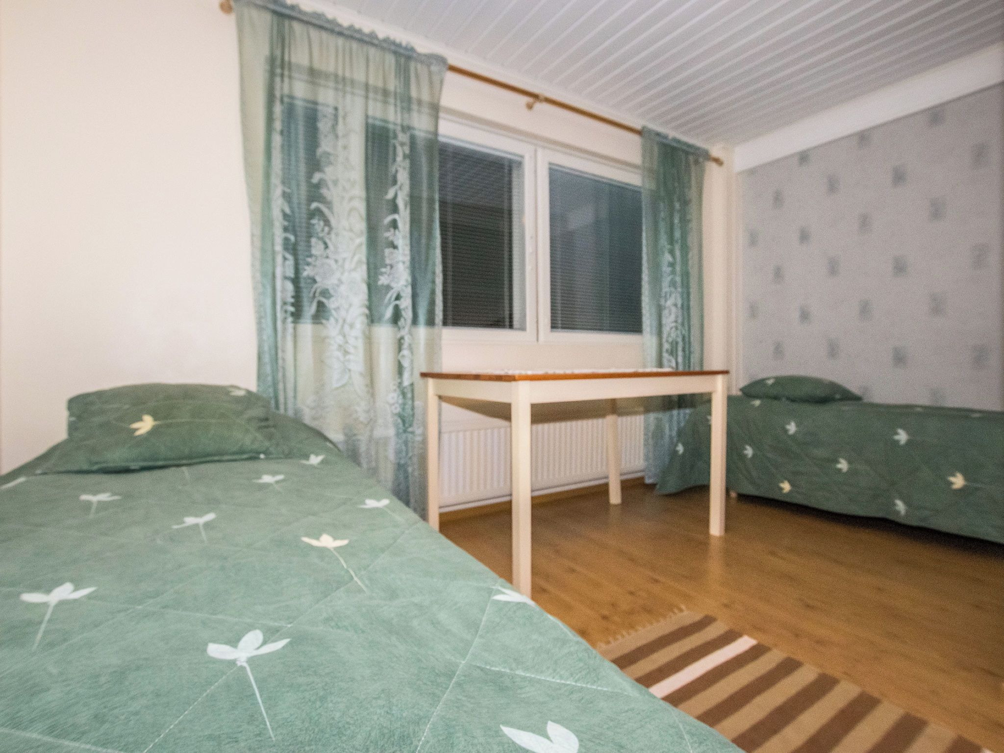 Habitació amb dos llits, una finestra amb cortines, un radiador i un terra de fusta.
