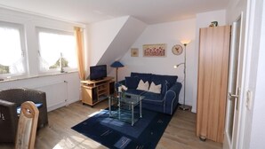 Un salon avec un canapé bleu, une table basse en verre, un fauteuil en osier, un meuble en bois et un téléviseur posé sur un meuble en bois.