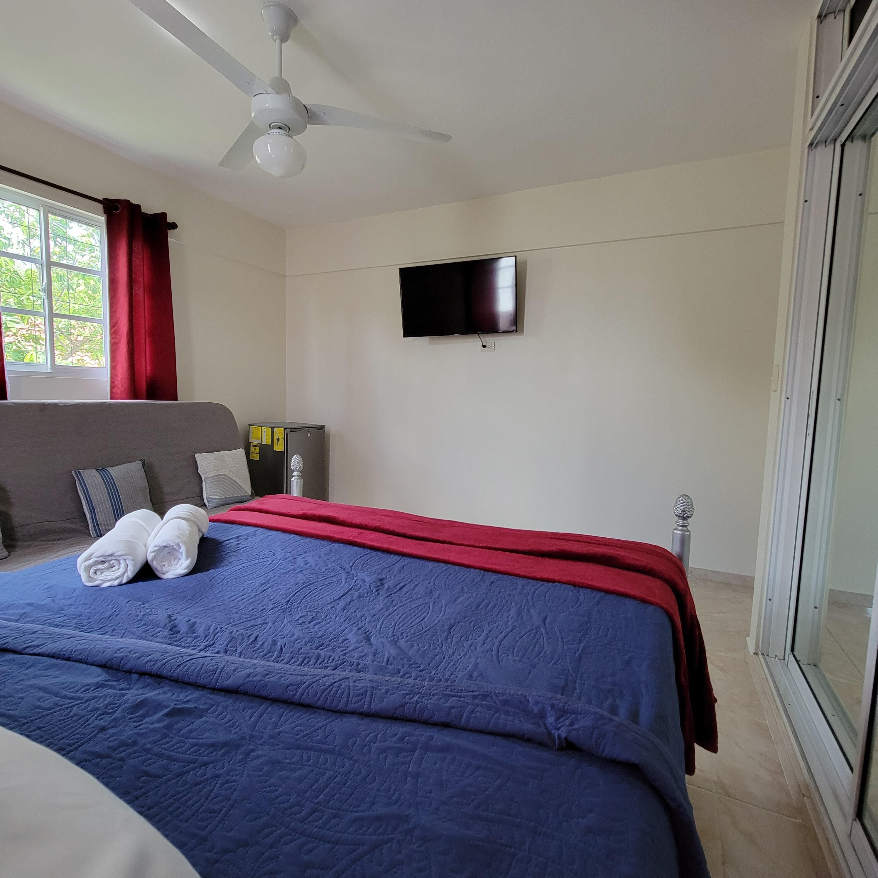 A hotel room with a bed, a ceiling fan, a TV on the wall, and a window with curtains.