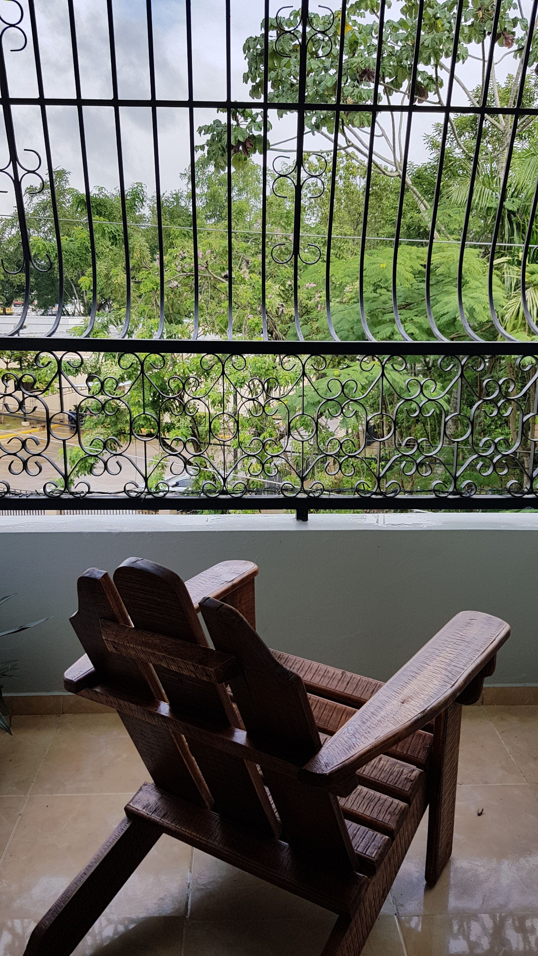 Una silla de madera en una habitación con vistas a un jardín a través de una reja. Se ven árboles y un edificio al fondo.