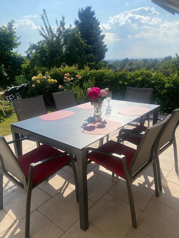 Eine Terrasse im Freien mit Tisch und Stühlen, umgeben von Grünpflanzen und einem wolkenlosen Himmel.