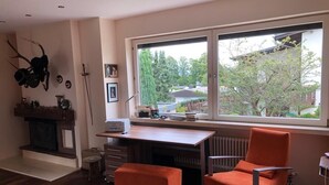 A modern living room with a wooden floor, a fireplace, a large window with a view of trees and houses, a desk with a computer, and two orange chairs.