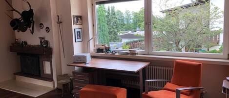 A modern living room with a wooden floor, a fireplace, a large window with a view of trees and houses, a desk with a computer, and two orange chairs.