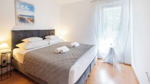 Een netjes opgemaakt bed met een grijs hoofdeinde, witte kussens en opgerolde handdoeken. Er staat een klein nachtkastje met een lamp en een bloempot. Aan de muur boven het bed hangt een schilderij.
