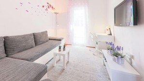 Een moderne woonkamer met een grijze bank, een witte salontafel, een flatscreen-tv aan de muur en een klein wit bureau met een potplant.