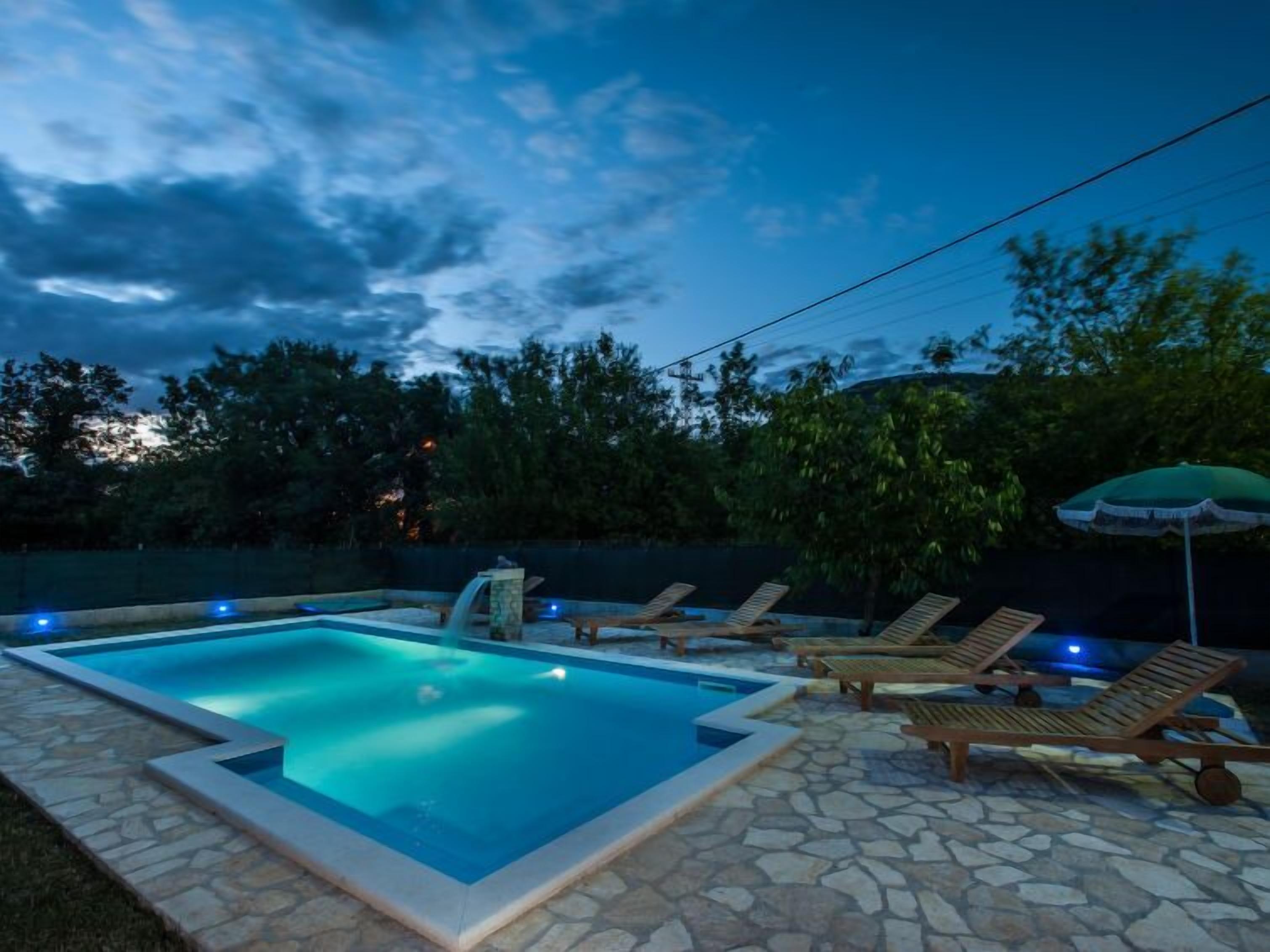 Una piscina exterior con borde de piedra, rodeada de tumbonas y una sombrilla.