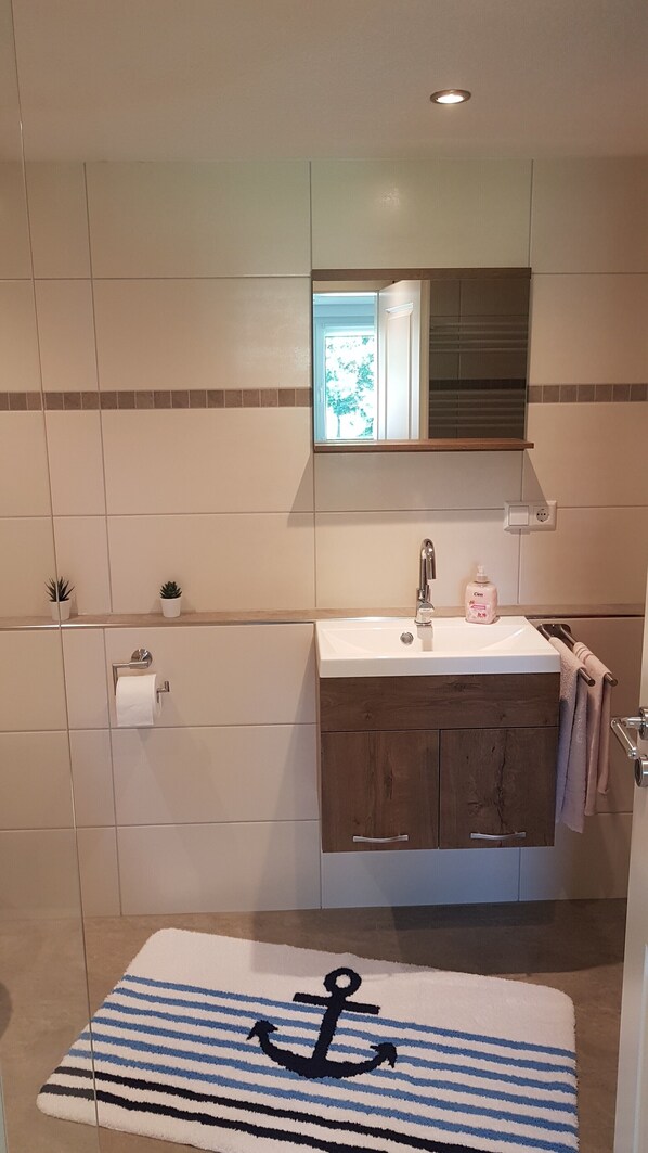 Un baño con un lavamanos blanco, armarios de madera, un espejo y un felpudo rayado con un diseño de ancla.
