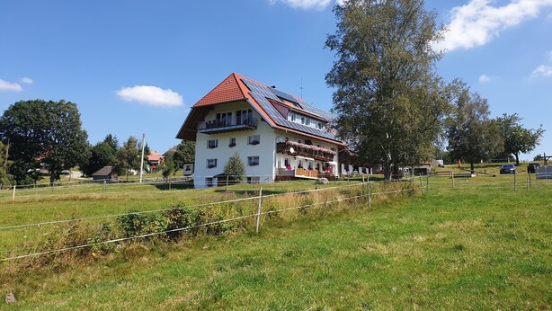 A two-story building with a red-tiled roof and solar panels, surrounded by a grassy field and trees.