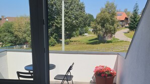 A balcony with a table and chairs, a potted plant, and a view of a green landscape with trees and a building.