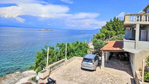 A coastal property with a stone-paved driveway, a parked car, and a view of the sea and distant islands.