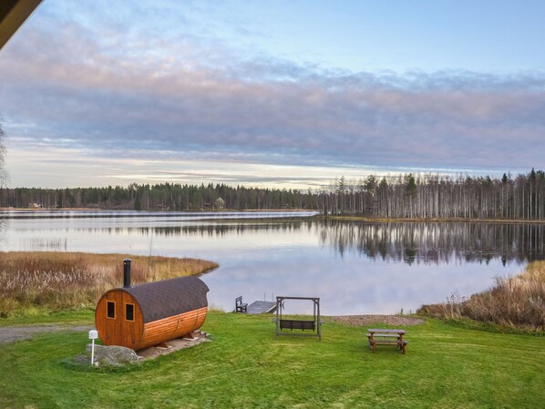 A cabin with a dock, a picnic table, and a bench by a calm lake with a forest in the background.