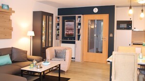 A modern living room with a sofa, armchair, and coffee table. There is a bookshelf, a dining area with chairs, and a kitchen with cabinets and a microwave. - Fantastic FH, on the dike, incl. Indoor pool use, play barn + lagoon free  (Butjadingen)