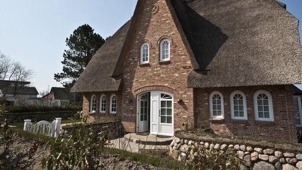 Ein Backsteinhaus mit einem steil geneigten Strohdach, einer mittleren Tür und zahlreichen Fenstern.