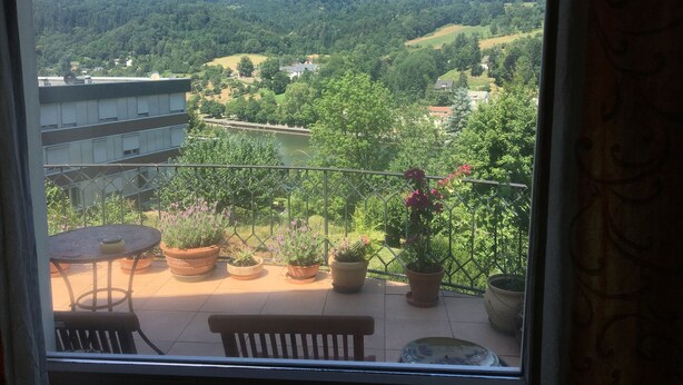 Un balcón con macetas y una mesa, con vistas a un río y un valle boscoso.