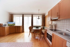 A kitchen with wooden cabinets, a dining table with chairs, a microwave, a sink, and a window with curtains.