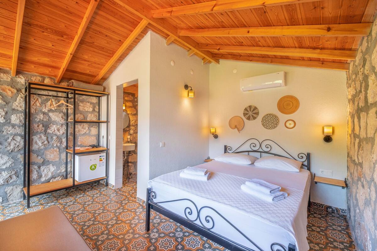 A bedroom with a stone wall, a bed with a metal frame, a wooden ceiling, and patterned floor tiles.