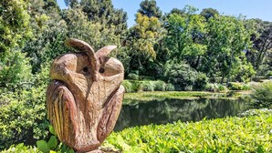 Una escultura de madera de un búho en un parque con un estanque y abundante follaje al fondo.