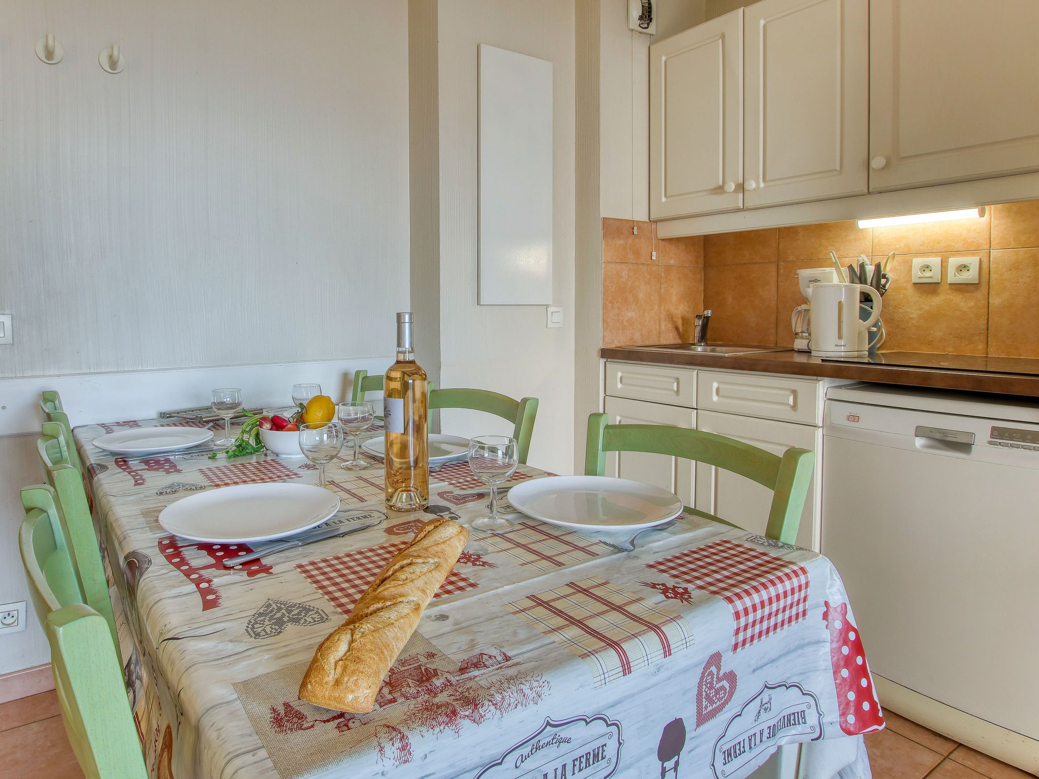 Un comedor con una mesa lista para cuatro personas, una botella de vino, pan y un cuenco con fruta.
