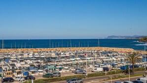 Un puerto deportivo con numerosas embarcaciones amarradas, una carretera junto al puerto y una vista despejada del océano y la costa distante.
