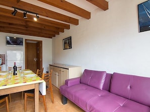 Una habitación con techo de madera, un sofá morado, una mesa de comedor con sillas, una pequeña cocina y un televisor montado en la pared.
