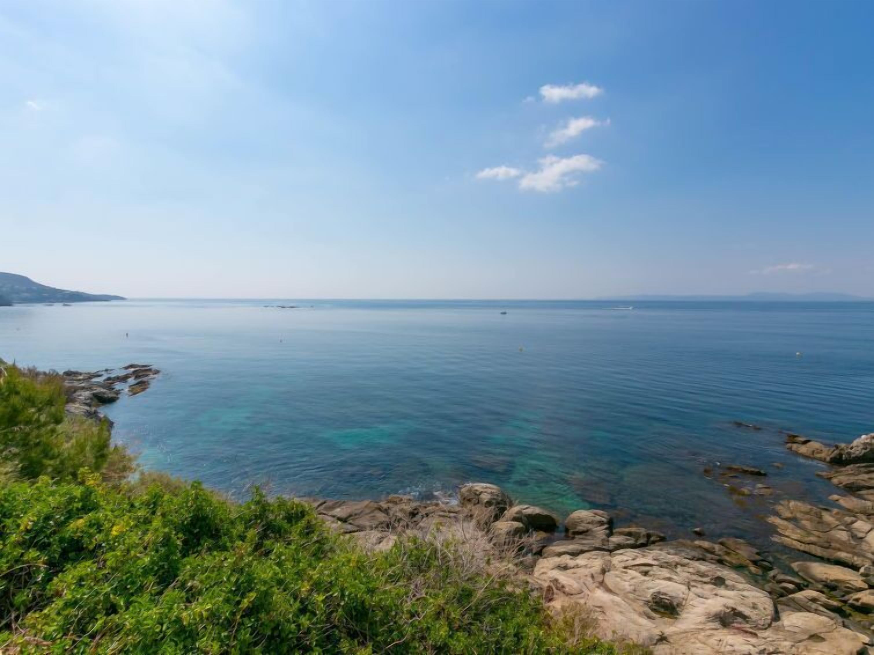 A coastal landscape with clear blue water, rocky shoreline, and distant landforms.