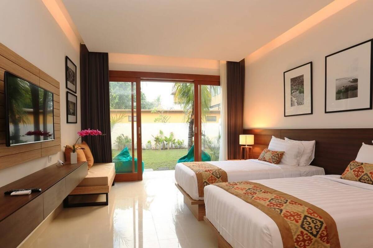 A hotel room with two beds, a TV, a desk, and a balcony with a view of greenery.