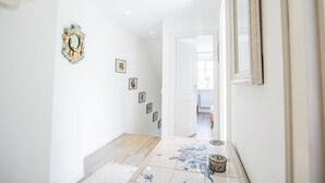 A hallway with white walls, a wooden floor, and a tiled floor with floral patterns. There are framed pictures on the wall and a decorative wreath on the left.