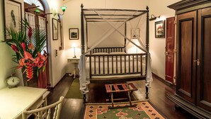 A traditional four-poster bed with a wooden frame, a bench at the foot of the bed, a small table with a lamp, and a large wooden door. - Royal Bar & Hotel (Kandy)