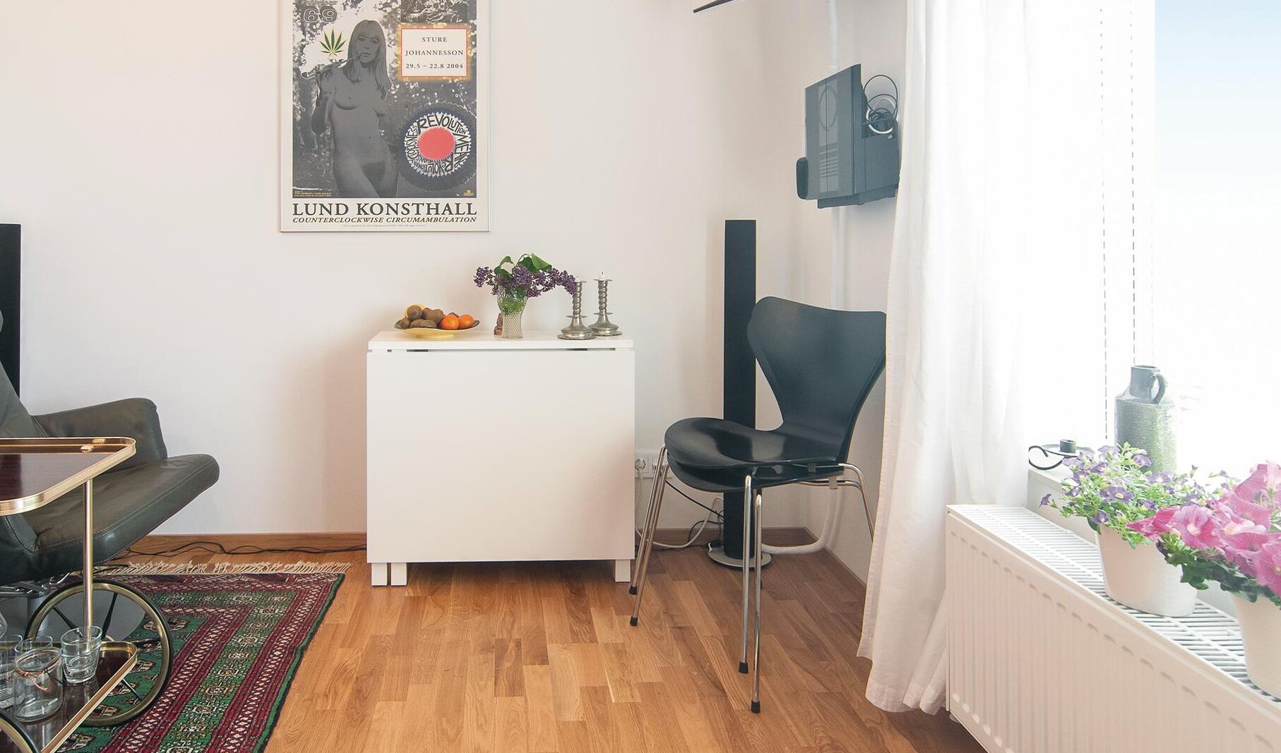 A modern living room with a white cabinet, a black chair, a small table with fruits and candles, a poster on the wall, and a window with plants.
