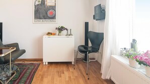 Una sala moderna con un mueble blanco, una silla negra, una mesita con frutas y velas, un cartel en la pared y una ventana con plantas.