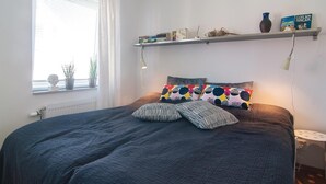 Un dormitorio con cama, ventana con persianas, radiador, estante con libros y un jarrón, y una lámpara de noche.
