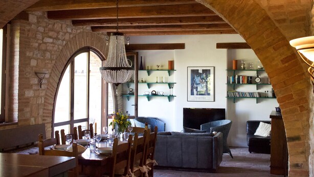 Un intérieur rustique avec des murs en pierre, des poutres en bois, un coin repas avec une table dressée pour quatre personnes et un coin salon confortable avec une télévision et des étagères.