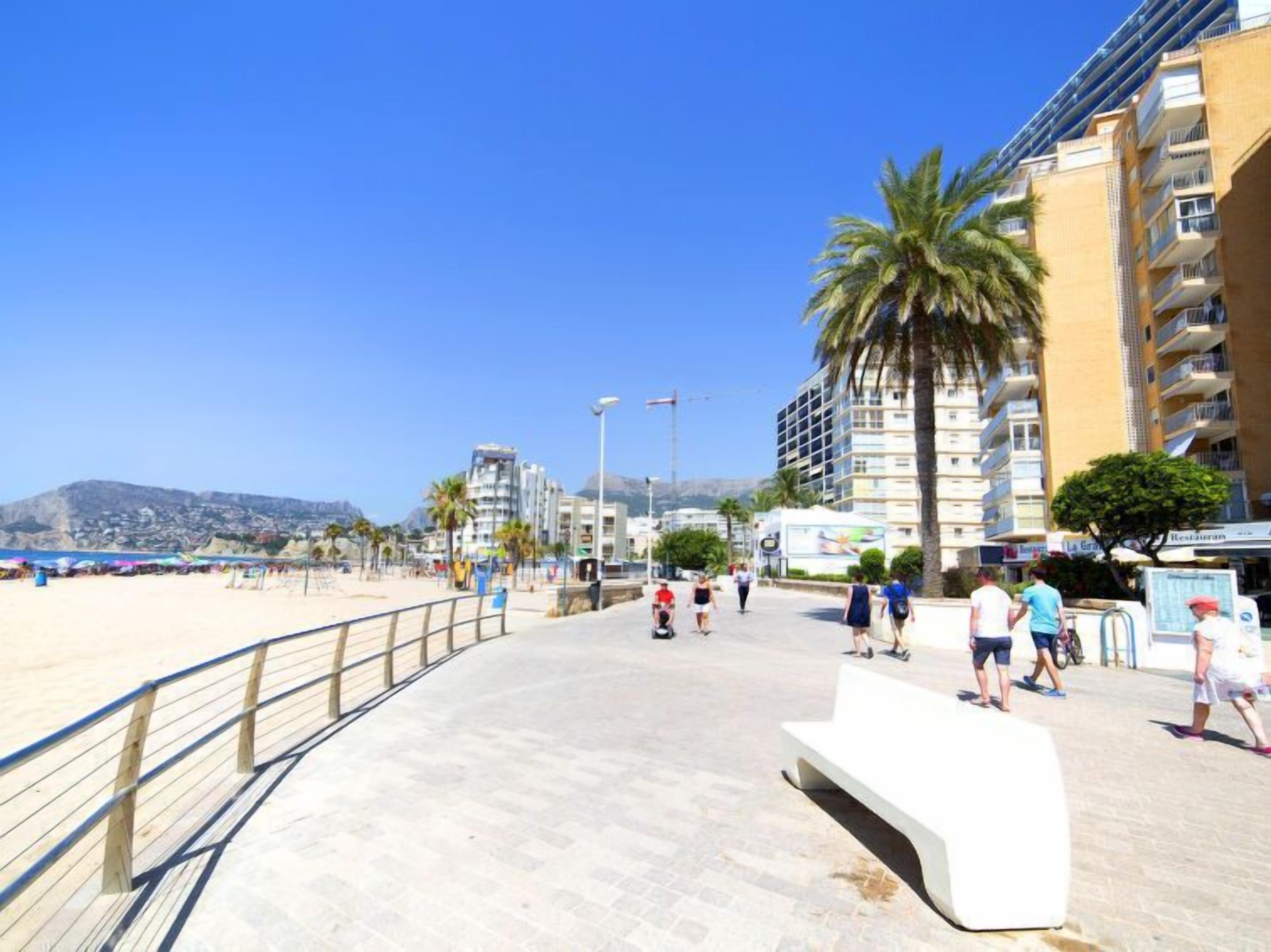 A coastal promenade with a sandy beach, palm trees, and modern buildings.