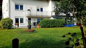 Un edificio de dos pisos con un balcón, césped cuidado y vegetación circundante.
