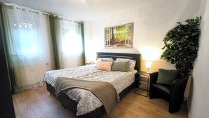 Un dormitorio con cama, mesita de noche, silla, planta en la pared y un cuadro.
