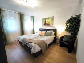 Ein Schlafzimmer mit einem Bett, einem Nachttisch, einem Sessel, einer Wandpflanze und einem Bild.