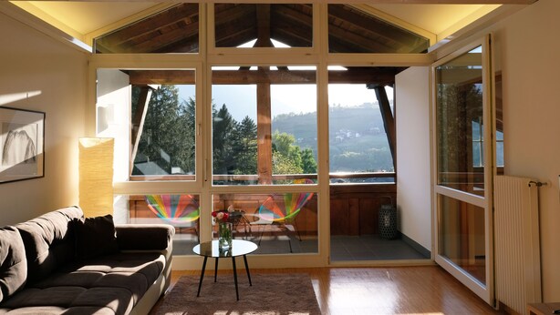 A room with a large window, a sofa, a small table with a vase, and a chair with a colorful backrest. - Refuge with a wonderful view (MERAN)