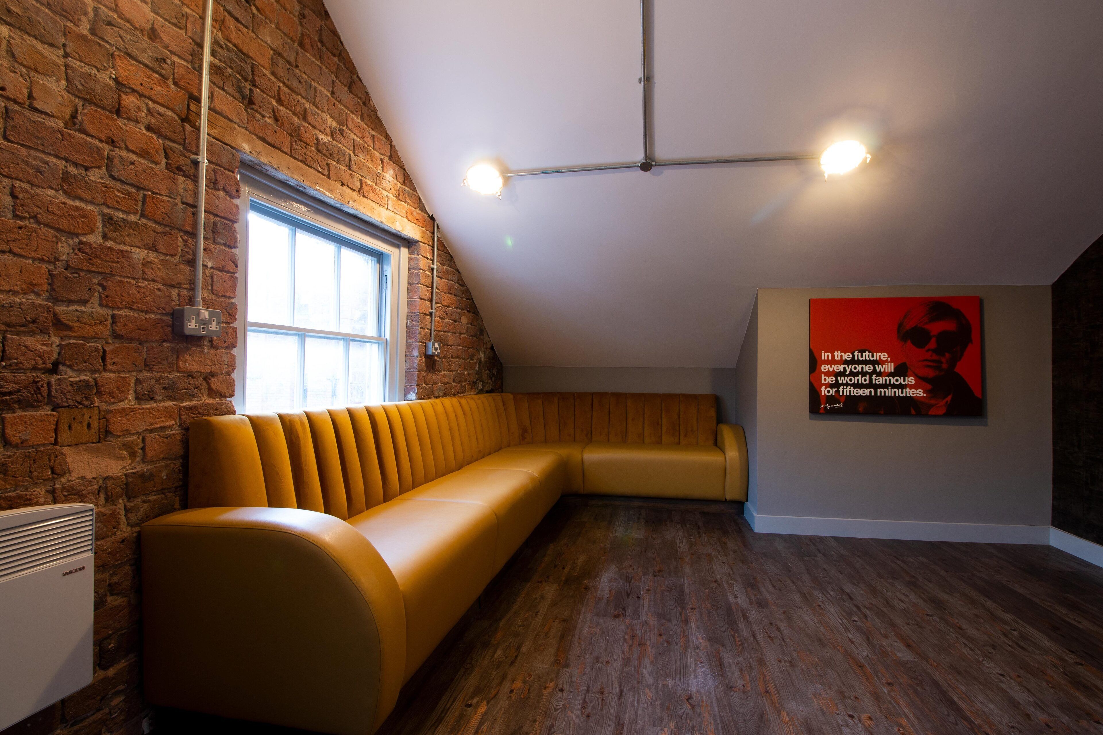 A room with a brick wall, a wooden floor, a yellow sofa, a window, and a poster on the wall.