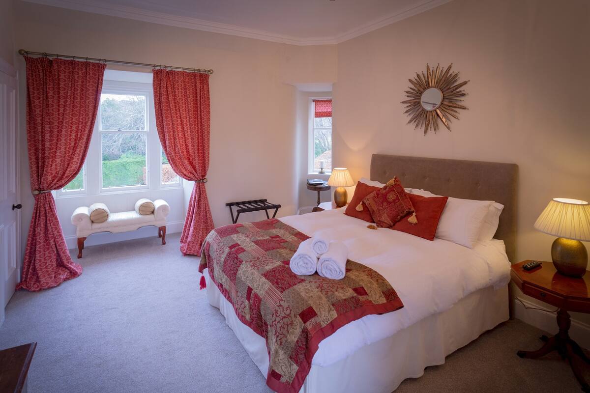 A neatly made bed with a red and beige quilt, a bench with cushions, a sunburst mirror, and a lamp on a bedside table.