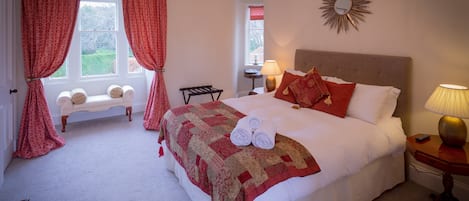 A neatly made bed with a red and beige quilt, a bench with cushions, a sunburst mirror, and a lamp on a bedside table.
