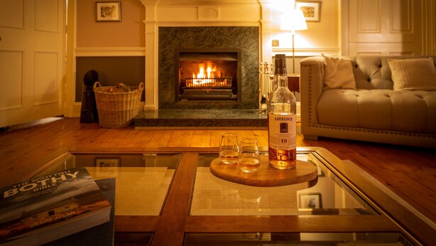 A cozy living room with a fireplace, a sofa, a glass coffee table with a bottle and glasses, and a bookshelf. - Craigellachie Lodge (Aberlour)