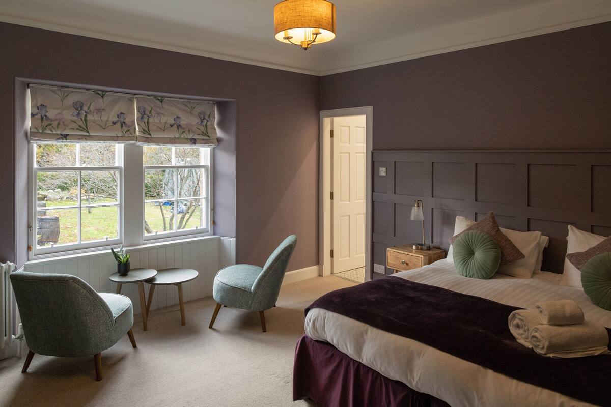 A bedroom with a large bed, two chairs, a small table, and a window with a view of the outdoors.