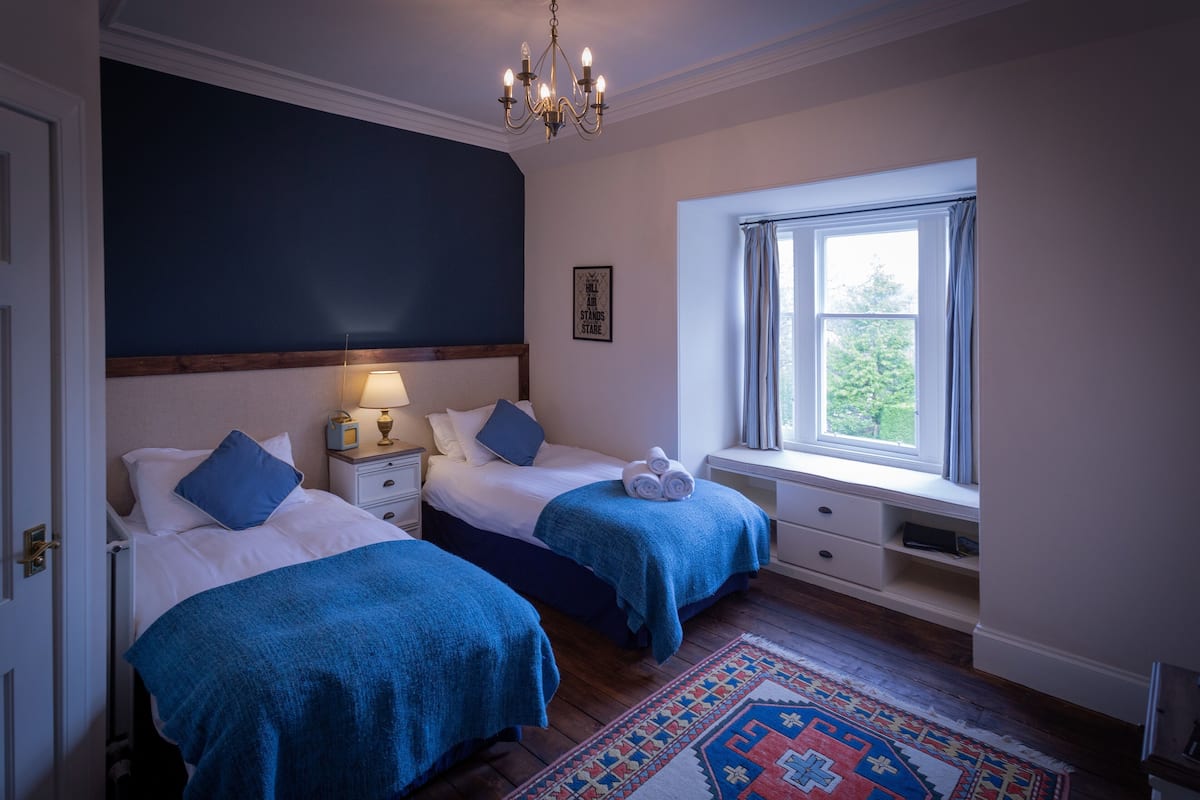A bedroom with two beds, a window with curtains, a patterned rug, and a chandelier.
