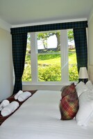 Una habitación de hotel con una cama grande, cortinas a cuadros, vistas a un paisaje verde y una lámpara en la mesita de noche.
